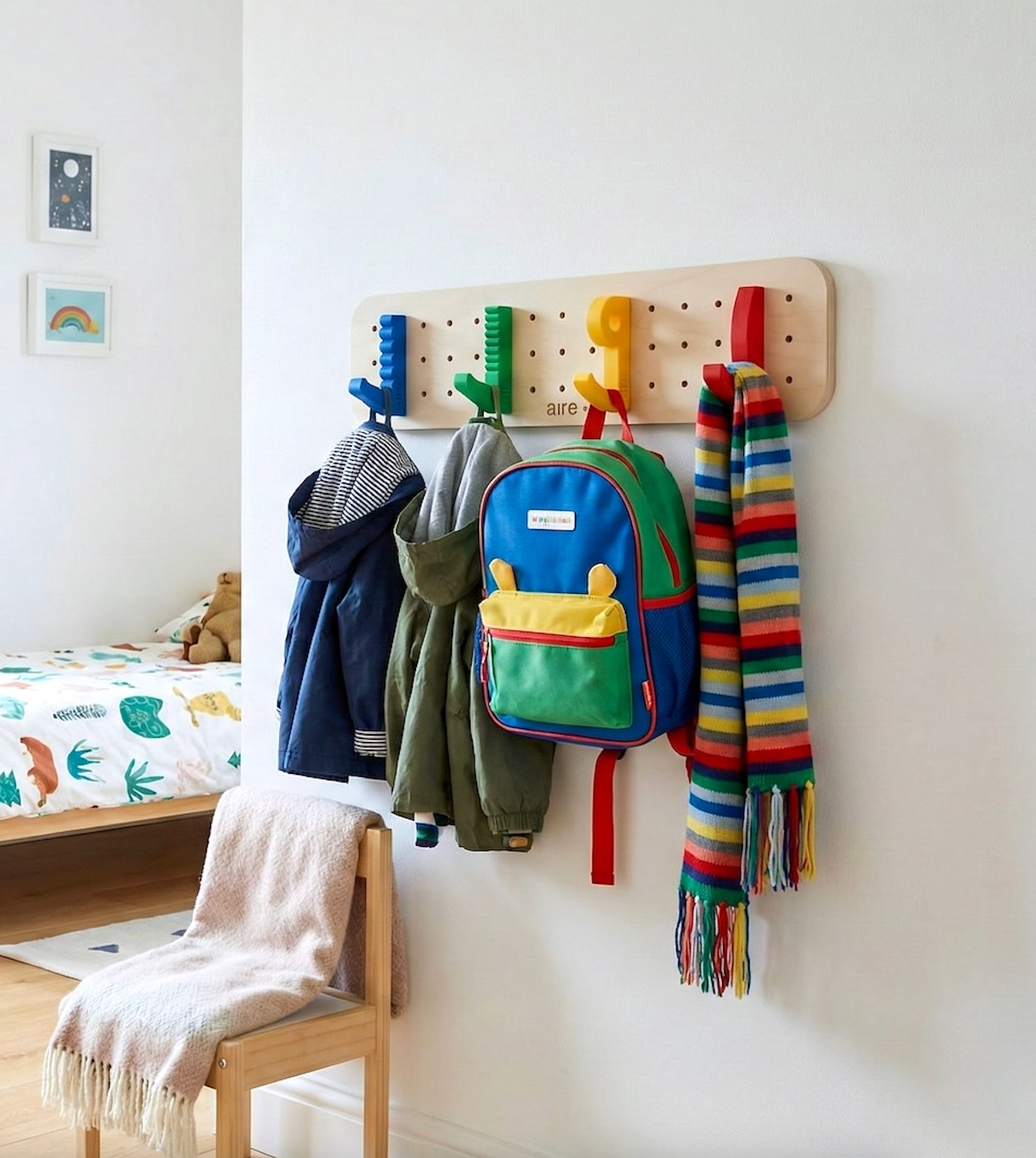 Wooden pegboard wall organizer with colorful hooks holding children’s coats, backpack, and striped scarf in a bedroom.