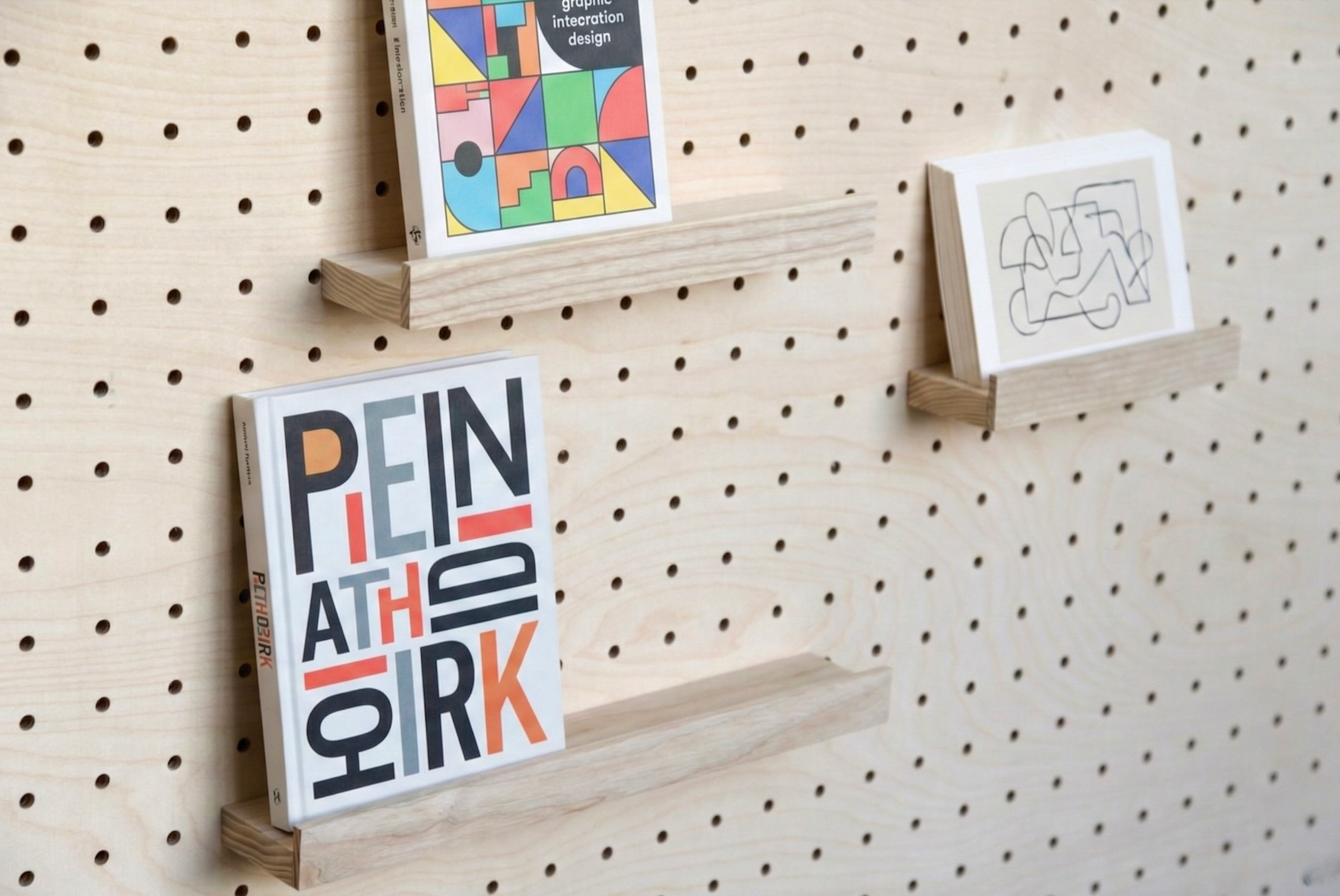 Minimalist wooden modular pegboard shelves holding books and artwork against a light pegboard wall.