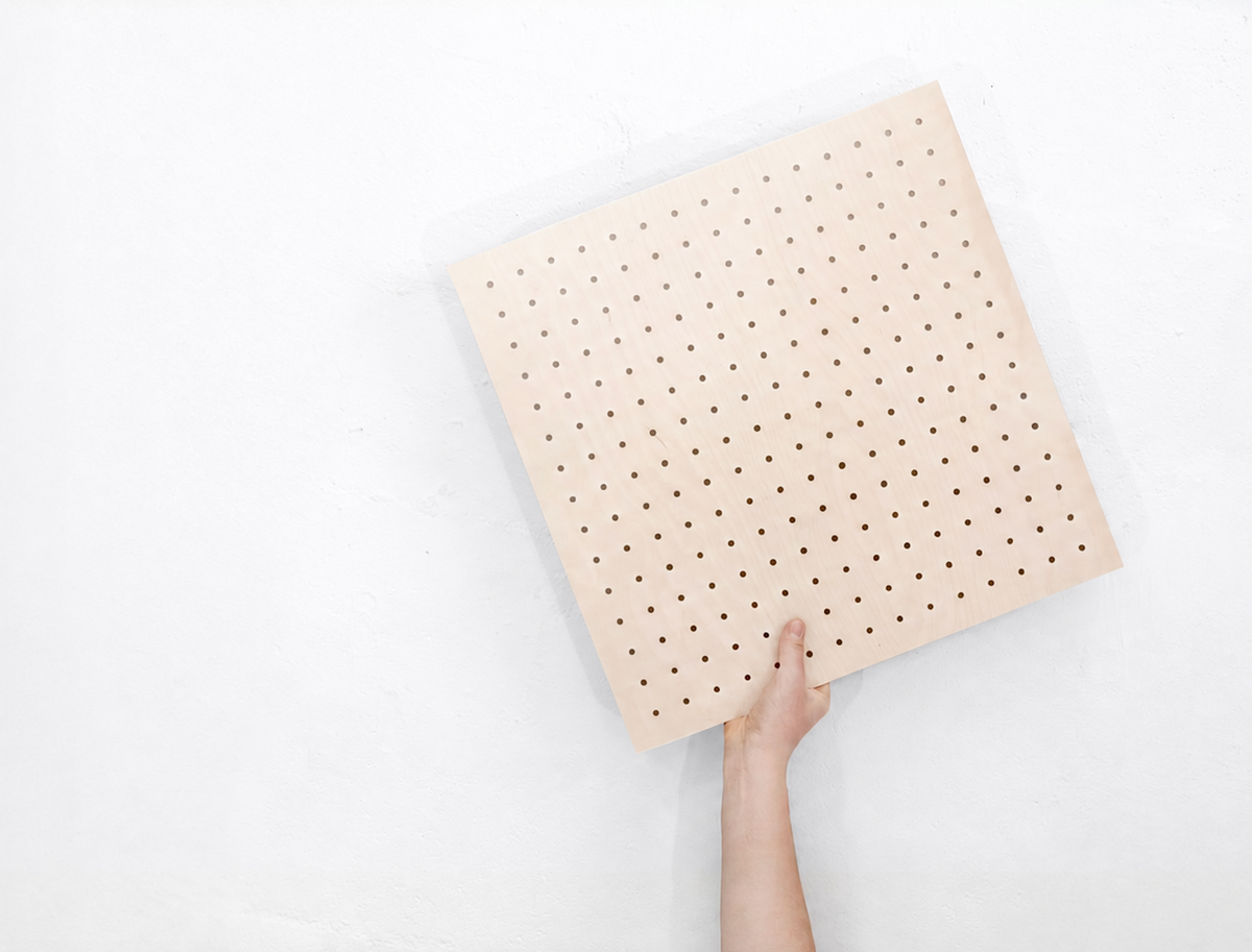 Pannello forato in legno di betulla 48 x 48 cm tenuto in mano davanti a una parete bianca