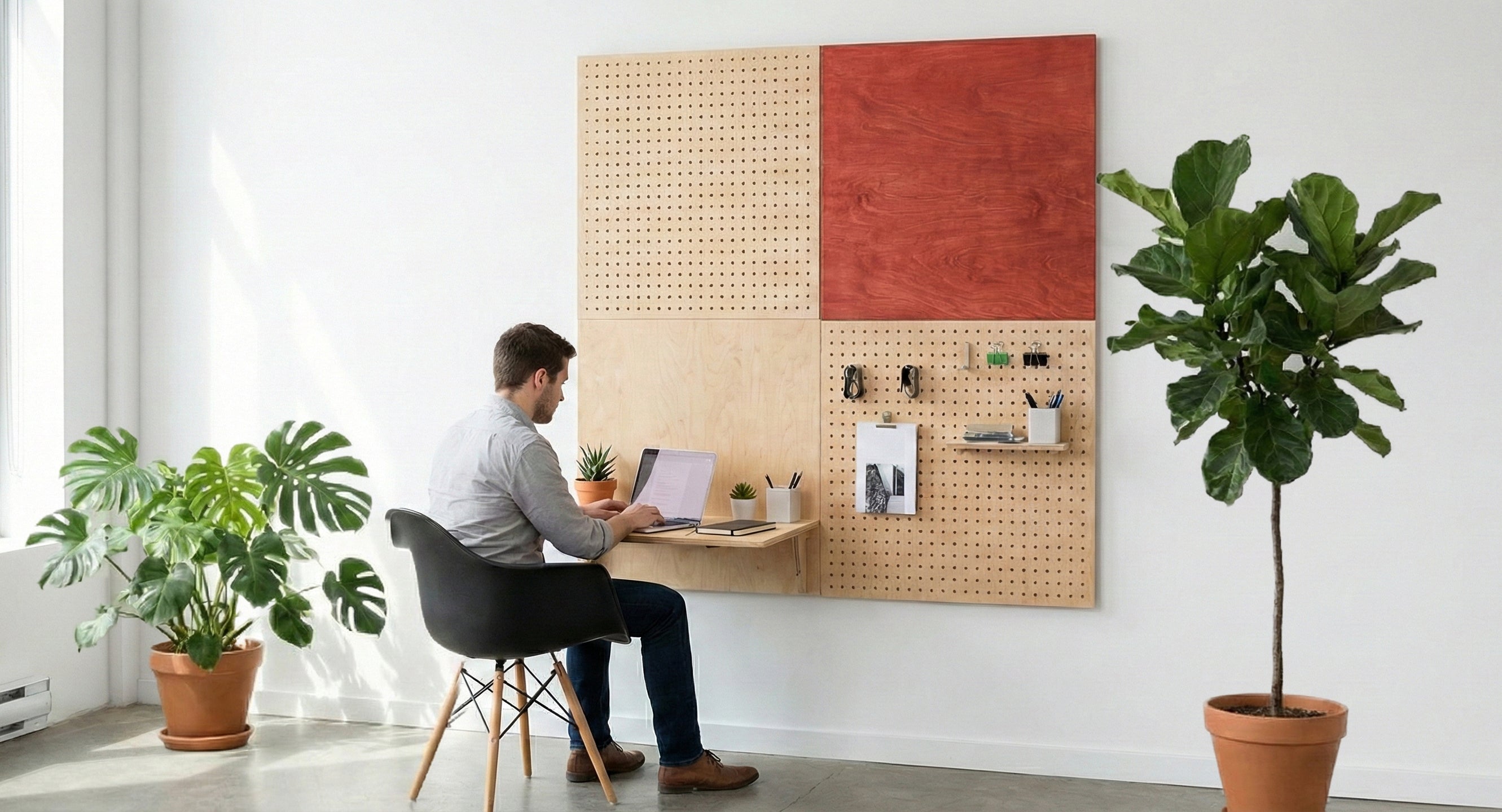 Man sitting at a chair working on laptop with modular pegboard shelf and wall organizer mounted on white wall nearby